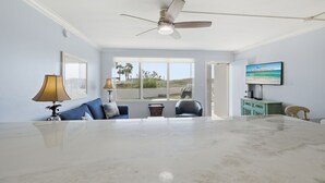 A spacious room with a large marble table, a ceiling fan, a TV, and a view of palm trees through the window.