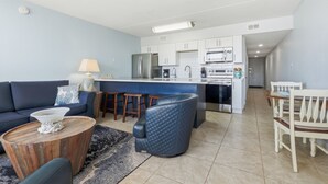 A modern living room with a sofa, coffee table, and a dining area with chairs. - Island Surf #8  1st Floor + Poolside + Beachfront + Beach Service! (Fort Walton Beach)