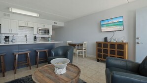 A modern kitchen with white cabinets, a microwave, and a coffee maker. There is a dining area with a table and chairs, a leather couch, and a television mounted on the wall. - Island Surf #8  1st Floor + Poolside + Beachfront + Beach Service! (Fort Walton Beach)