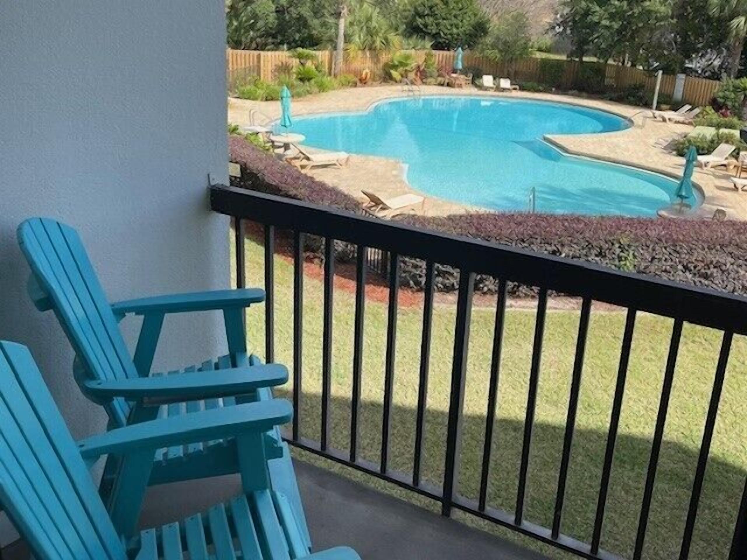 A balcony with a view of a swimming pool, surrounded by a wooden fence and greenery.