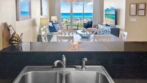 A kitchen with a double sink, a dining area with a table and chairs, and a view of the ocean through large windows.