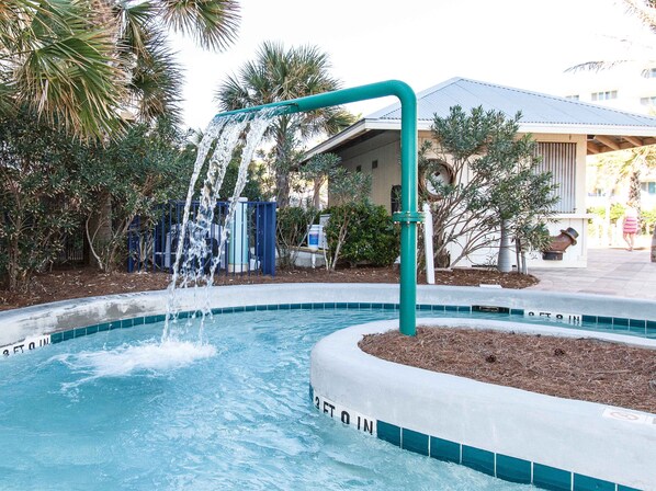 Une piscine agrémentée d’un élément décoratif aquatique, entourée de palmiers et avec un bâtiment visible au loin.