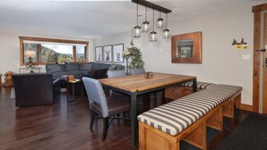 Uma sala de estar acolhedora com uma mesa de jantar de madeira, um banco e um sofá. Há luminárias penduradas sobre a mesa e uma janela grande com vista para árvores.