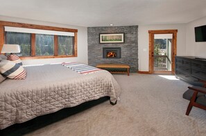 A bedroom with a large bed, a fireplace, a bench, a TV, and a balcony door.