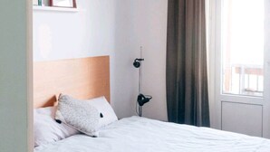 Een slaapkamer met een houten hoofdeinde, een bed met wit beddengoed, een nachtkastje met een lamp en een raam met gordijnen.