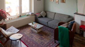 Een woonkamer met een grijze bank, een tapij met dessin, een bijzettafeltje en een raam met jaloezieën.