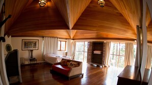 A spacious room with a wooden ceiling, a bed, a red sofa, and a large window offering a view of the outdoors. - Surya Pan Hotel e Eventos (Campos do Jordão)