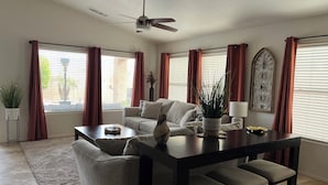 A living room with a sofa, a coffee table, a dining table with four chairs, a ceiling fan, and large windows with curtains. - 5 STAR Goodyear Spring Training Hiking Mountain Views Heated Pool Included (Goodyear)