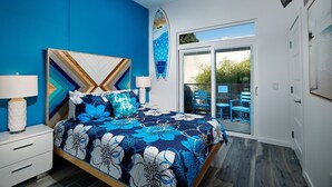 A bedroom with a bed, a nightstand, a lamp, a surfboard art piece, and a sliding glass door leading to an outdoor area with blue chairs.
