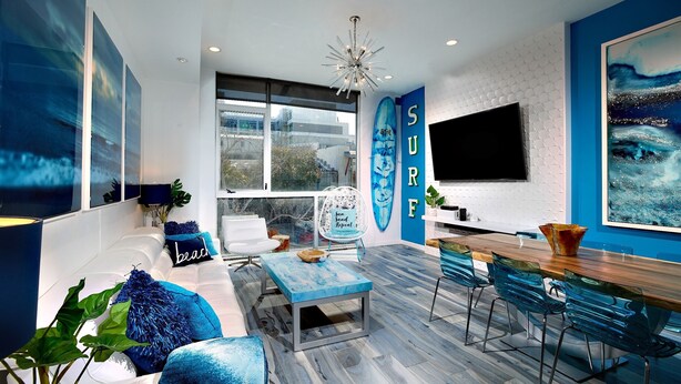A modern living room with a large window, a blue sofa, a coffee table, and a dining area with chairs and a wooden table.