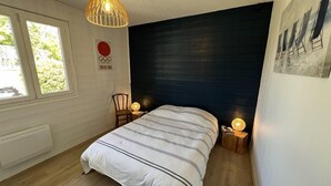 A bedroom with a bed, a chair, a nightstand, a hanging light fixture, a window, and a wall-mounted picture.