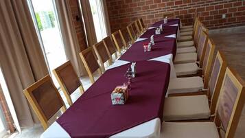 A long dining table with white tablecloths and wooden chairs, set against a brick wall with a TV mounted above.