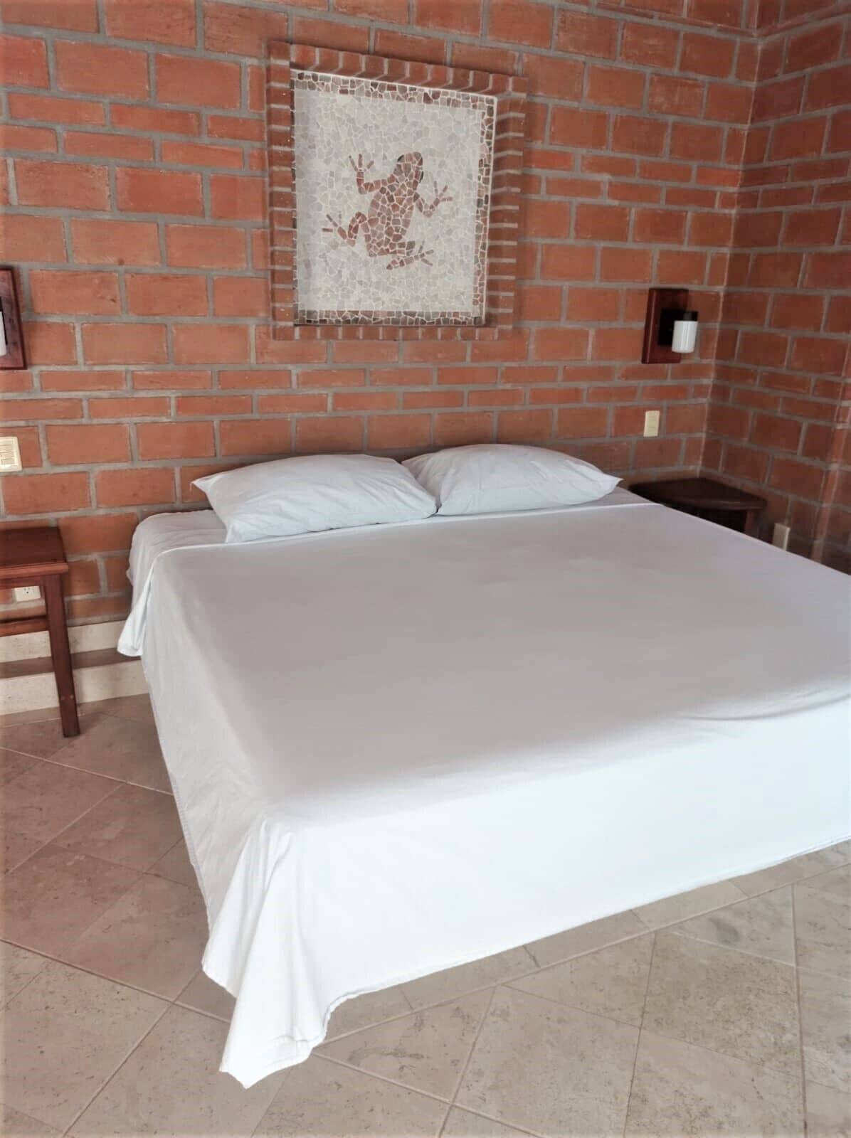 A room with a brick wall, a bed with white bedding, a small table, and a wall-mounted light fixture.