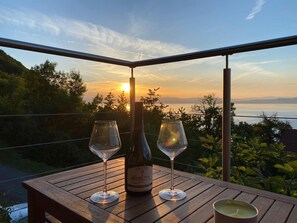 Een houten tafel met een wijnfles en twee wijnglazen op een balkon met uitzicht op een zonsondergang boven het water.
