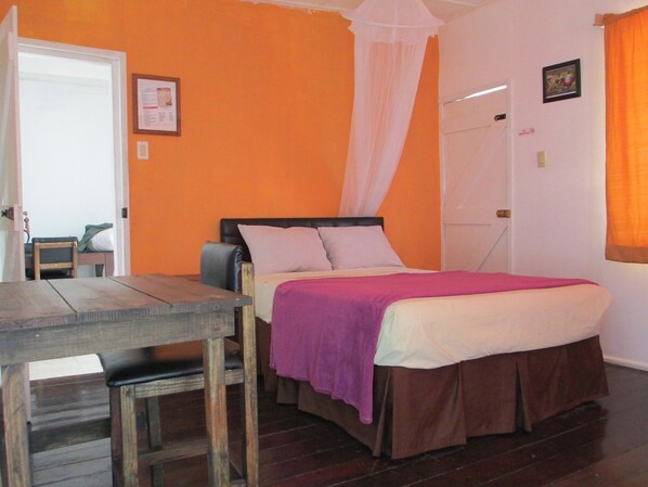 A bedroom with an orange wall, a bed with a brown skirt, a wooden table, and a chair.