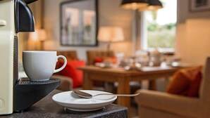Een keukenhoek met een koffieapparaat, een kop en een lepel op een dienblad. Er staat een eettafel met stoelen en een lamp op de achtergrond.