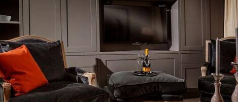 A dark-themed living room with a sofa, ottoman, and a glass table with a book and candle.
