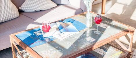 A glass-top coffee table with magazines and two wine glasses, placed on a rug in a living room with a sofa and cushions.
