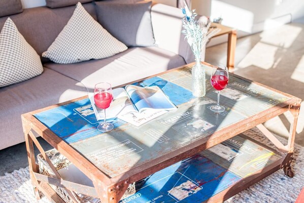 A glass-top coffee table with magazines and two wine glasses, placed on a rug in a living room with a sofa and cushions.