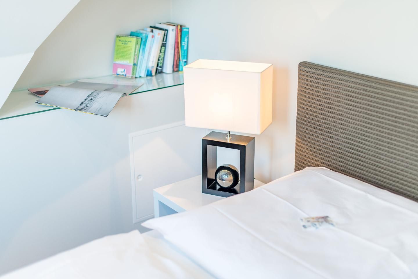 Una cama bien hecha con sábanas blancas, una lámpara de noche y una estantería con libros y una revista.
