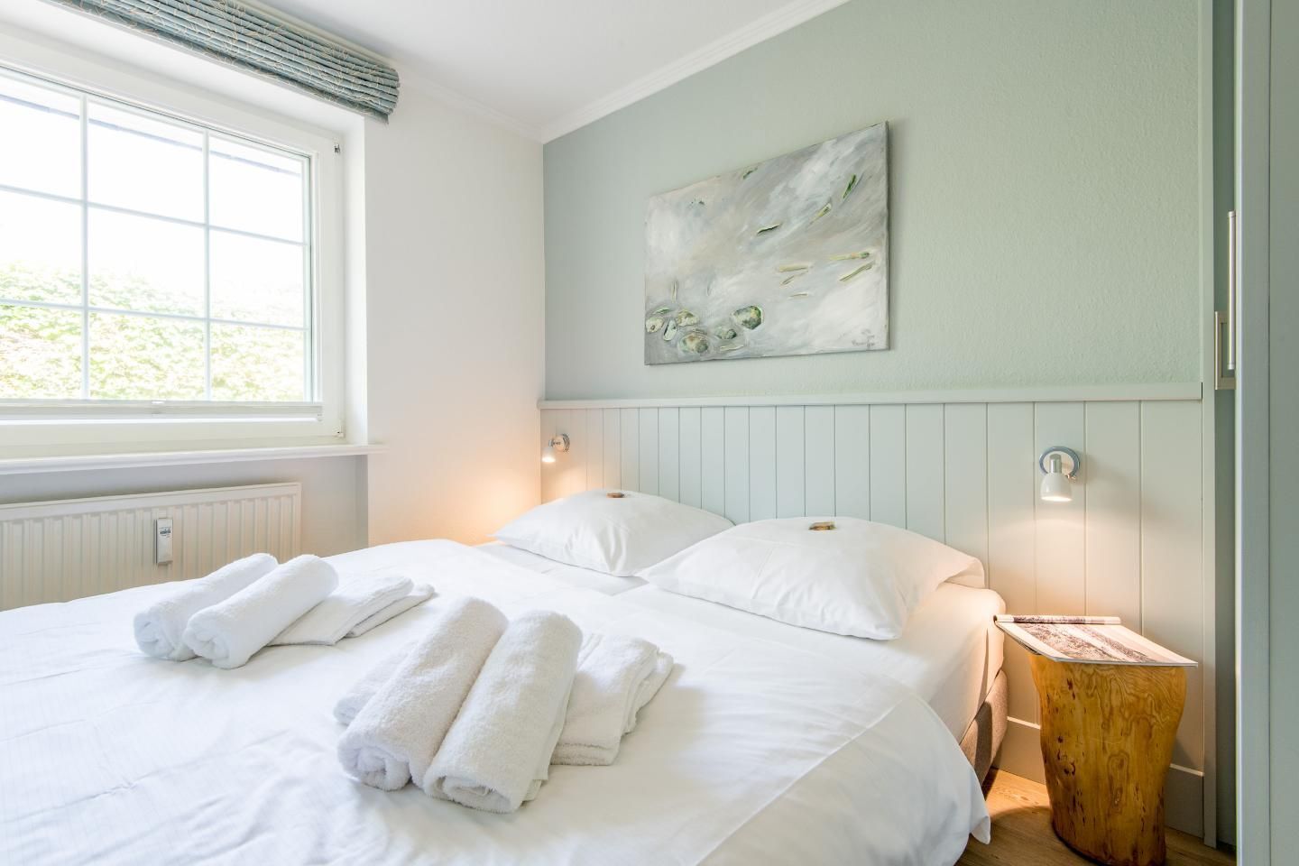 A neatly made bed with folded towels, a wooden side table, and a wall-mounted abstract painting.