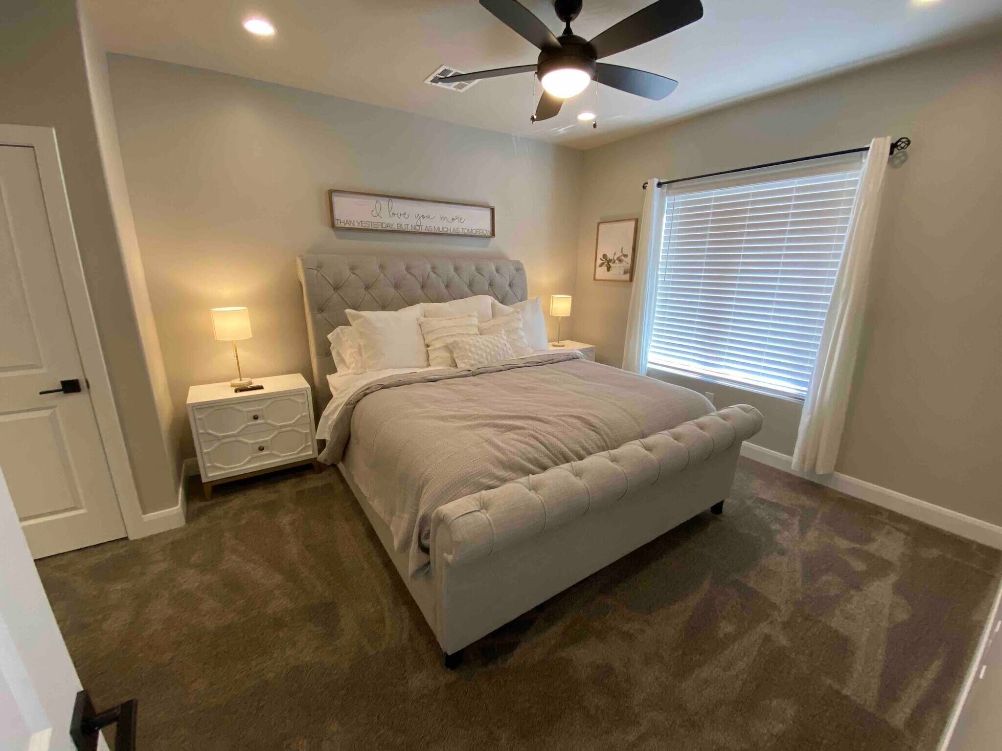 A bedroom with a tufted headboard, a large bed, two bedside tables with lamps, a ceiling fan, and a window with blinds.