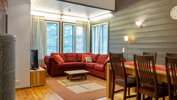 Een gezellige woonkamer met een rode bank, een houten eettafel en stoelen. Er staat een televisie op een dressoir, een open haard en grote ramen met gordijnen.