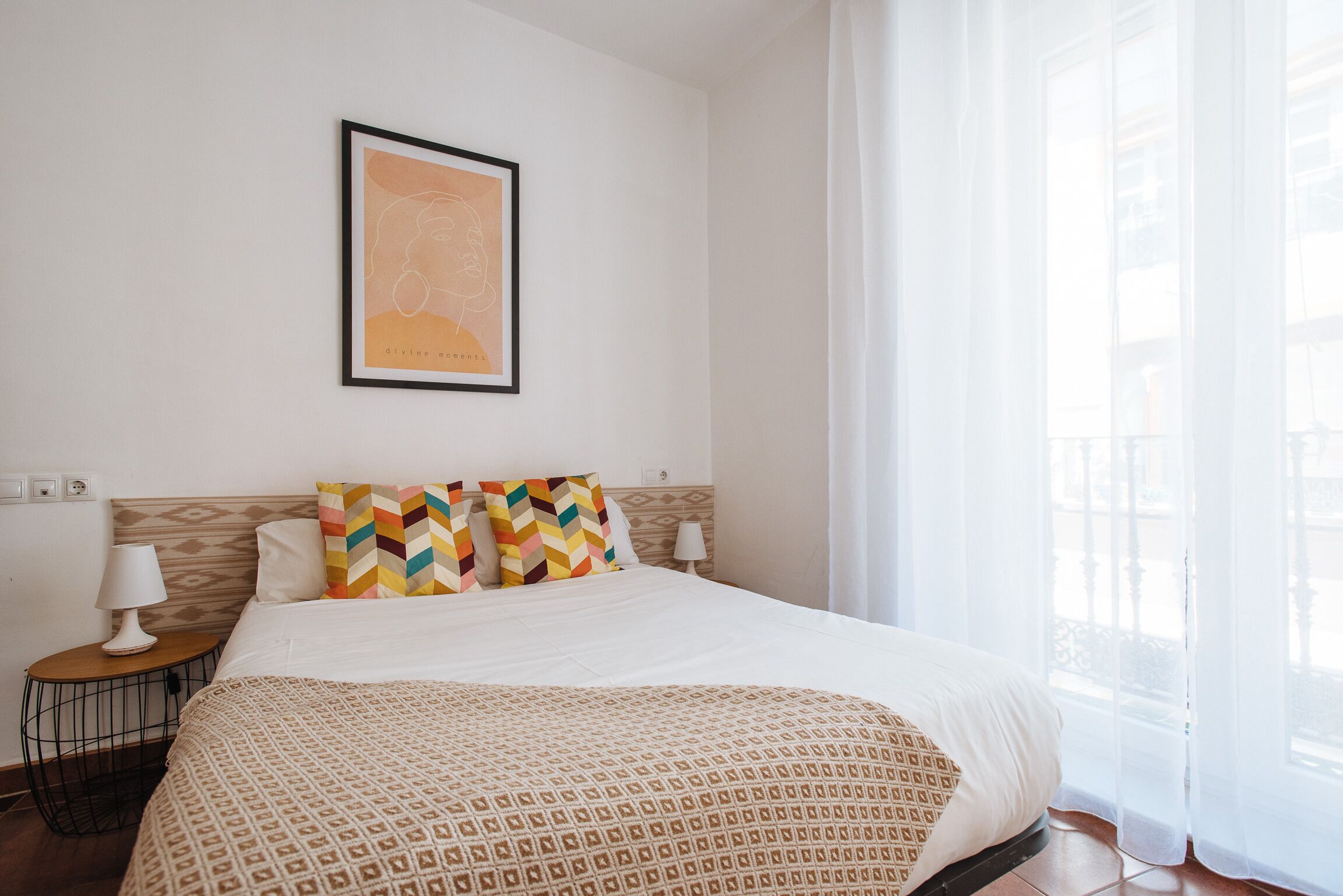 A hotel room with a bed, a nightstand, a lamp, a framed picture on the wall, and a window with sheer curtains.
