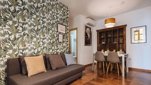 A living room with a sofa, a dining table with chairs, a wooden cabinet, and a wall with a leafy wallpaper. - Homey Seville. Bright Apartment in Santa Cruz (Sleeps 6; Parking Optional) (Sevilla)