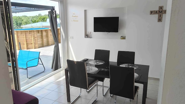 A dining area with a TV mounted on the wall, a table with four chairs, and a view of a wooden deck outside. - Sunbeam Bottom of villa T3 beach 2km away  (Sainte-Anne)