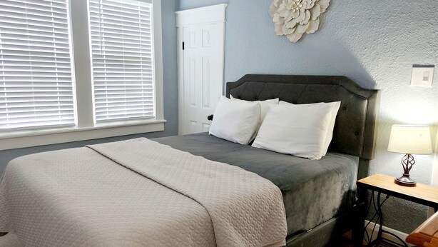 A bedroom with a bed, white blinds, a white door, a decorative wall piece, and a bedside table with a lamp. - Lovely One Bedroom Home (Lakeland)