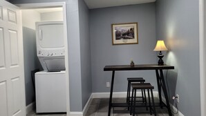 A laundry room with a washing machine, a table with a lamp, and a framed picture on the wall. - Lovely One Bedroom Home (Lakeland)