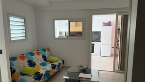 Une pièce comprenant un canapé, une petite table, un ventilateur de plafond, une fenêtre et donnant sur une autre pièce.