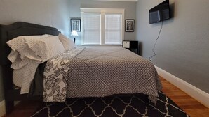 A bedroom with a bed, a ceiling fan, a window with blinds, and a wall-mounted TV.