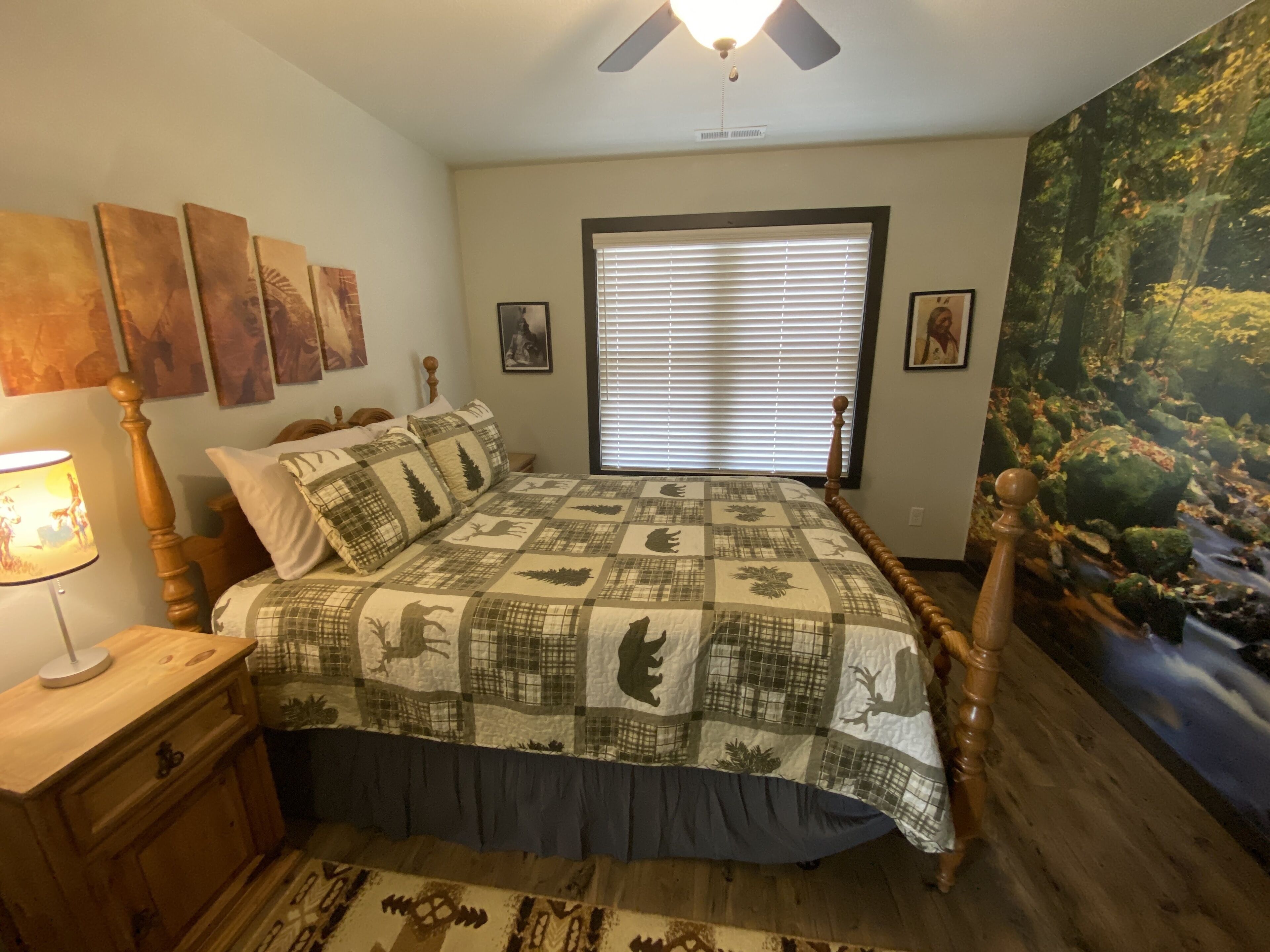 Un dormitorio con cama, mesita de noche, lámpara, ventilador de techo y un gran mural con motivos forestales.