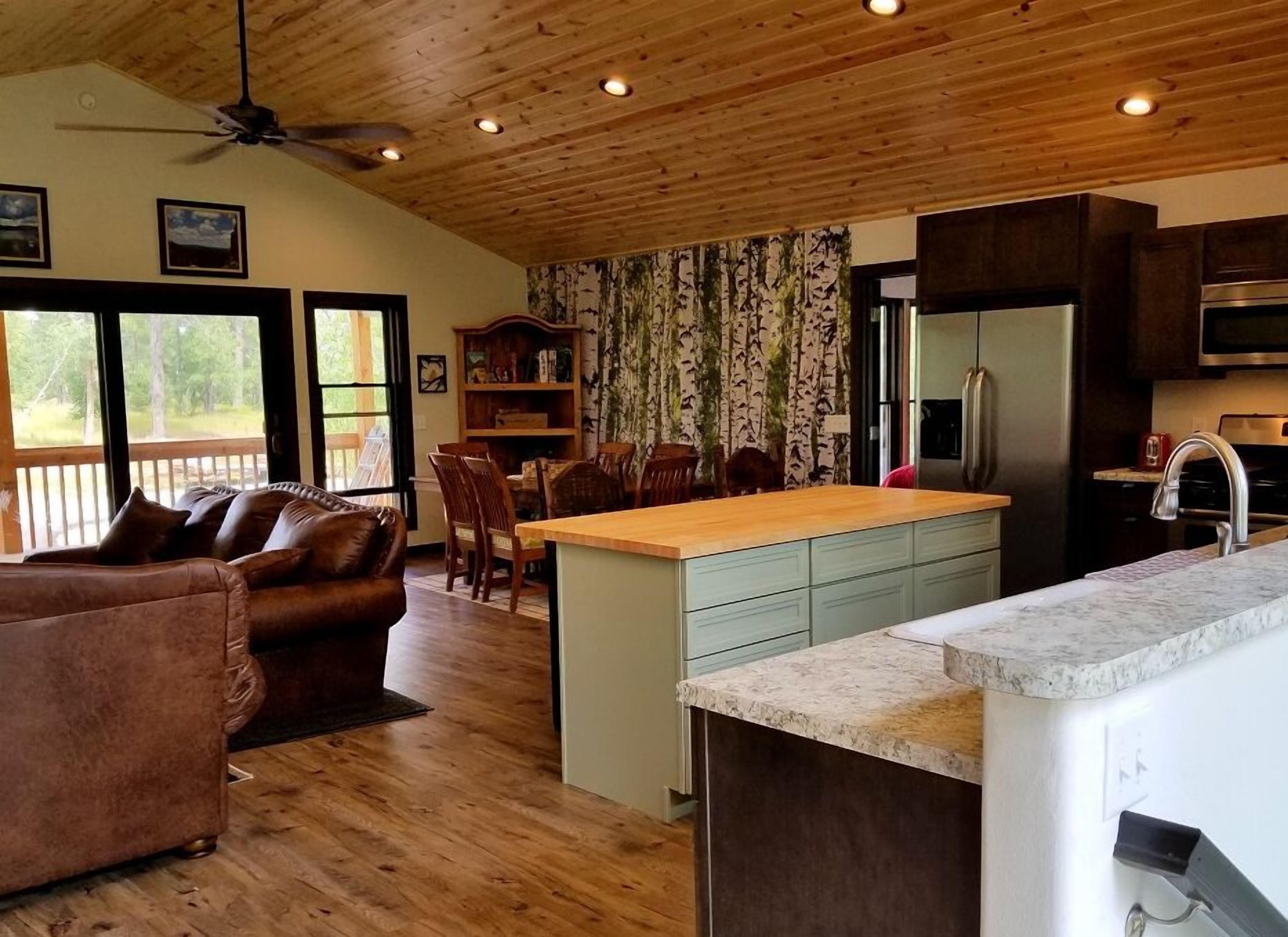 A cozy cabin interior with a wooden ceiling, a dining area, a kitchen with a refrigerator, and a large window overlooking a forest.