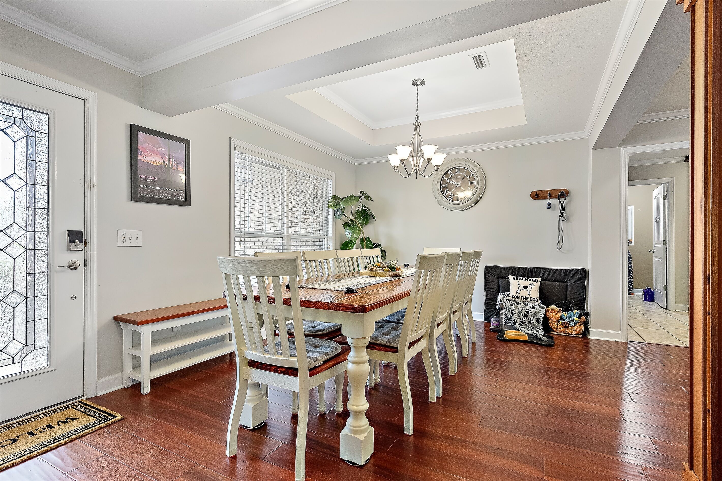 A dining area with a wooden table and white chairs, a chandelier, and a large window with blinds.