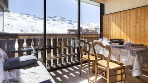 Une pièce chaleureuse avec un plafond en bois, une grande fenêtre donnant sur une montagne enneigée, une table à manger avec des chaises et un lit recouvert d’une courtepointe à motifs.