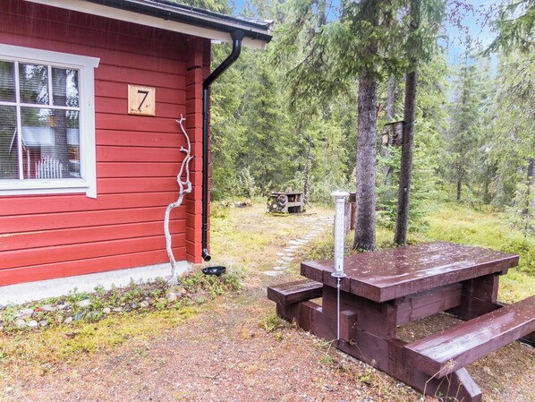 En rød hytte med et skilt med nummer 7, et picnicbord og et fuglefoderhus i et skovområde.