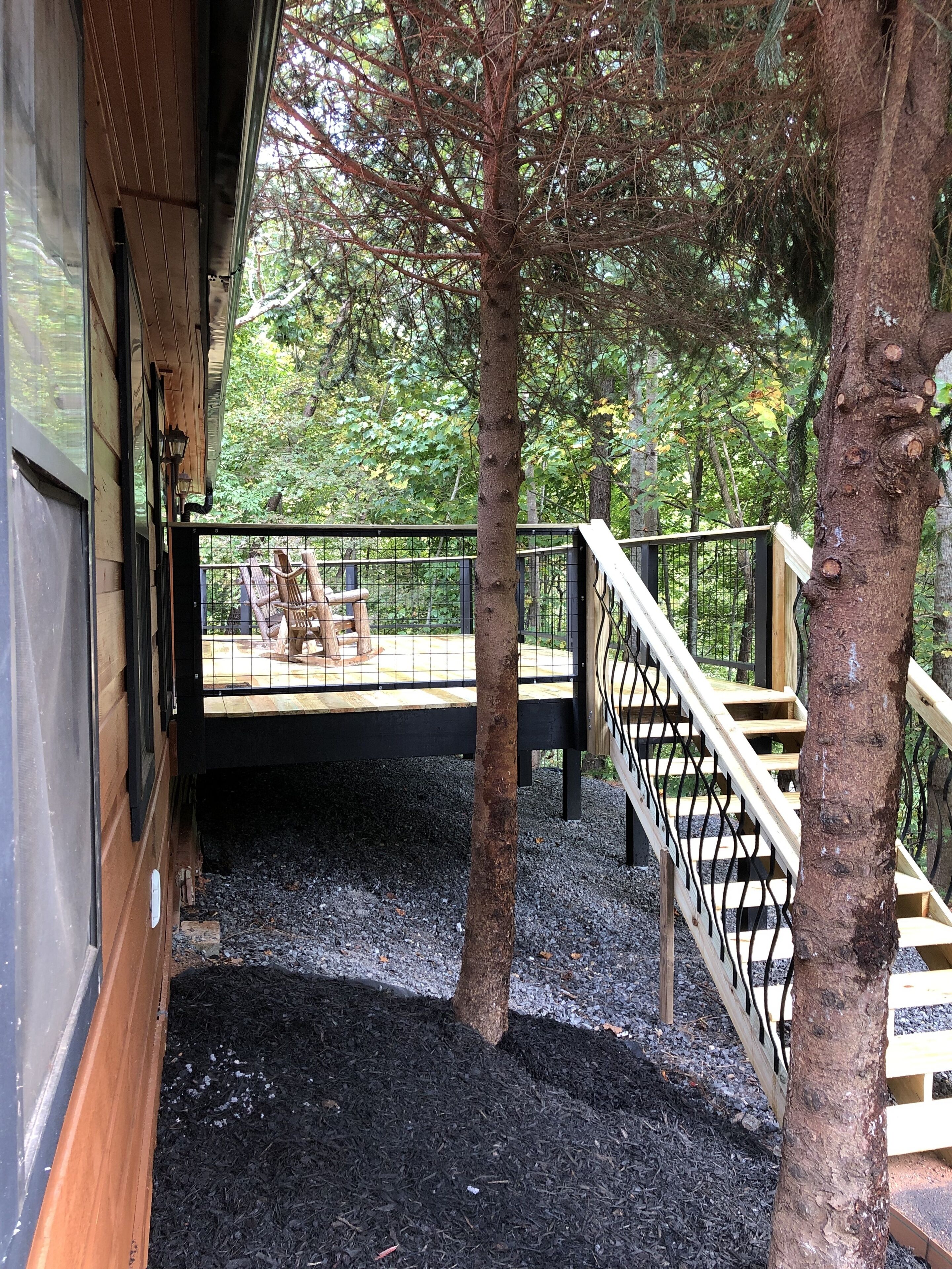 Una terraza de madera con barandilla negra, rodeada de árboles y un sendero de grava.