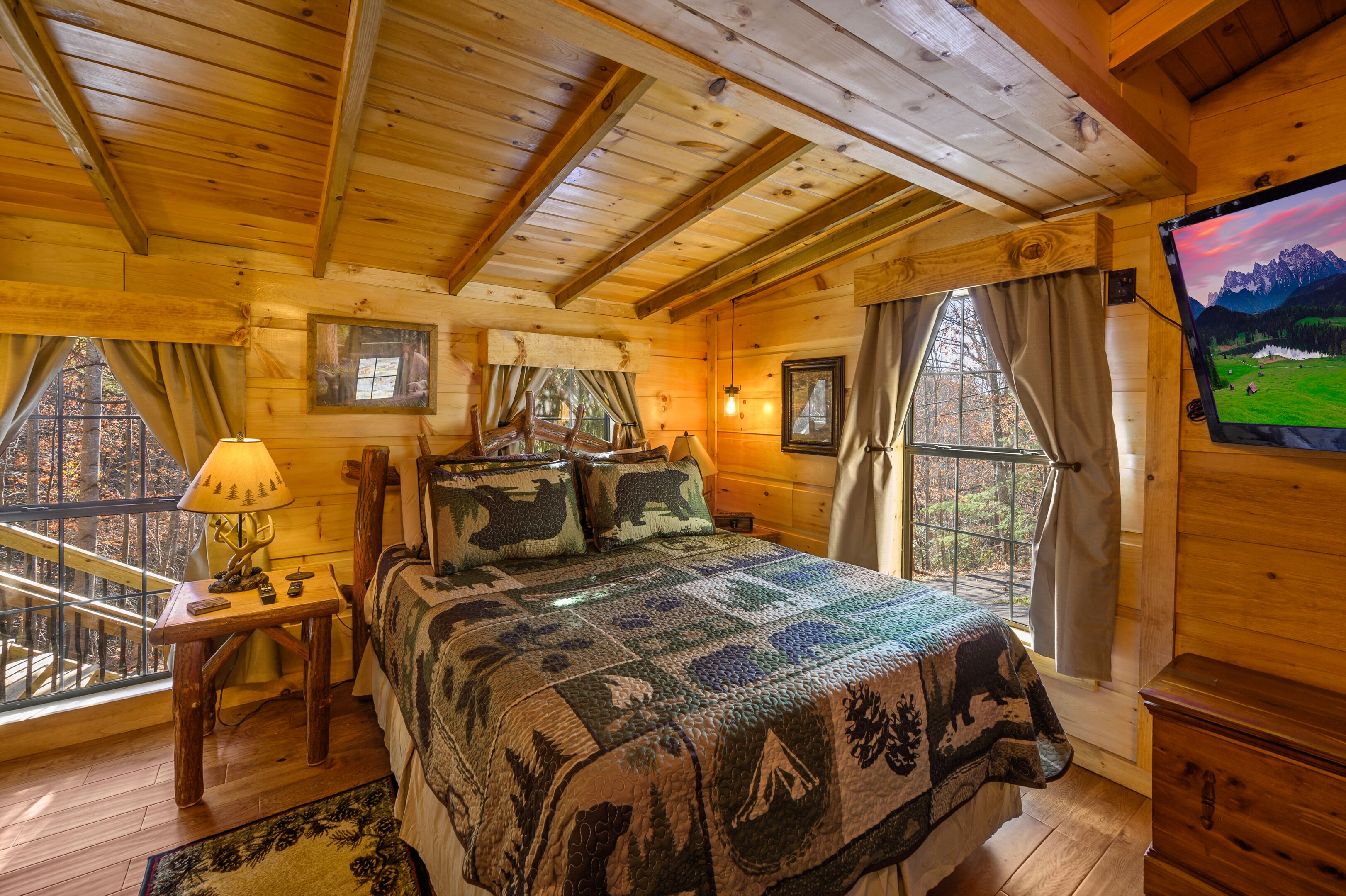 A cozy wooden cabin bedroom with a large bed, a small table with a lamp, and a TV on the wall.