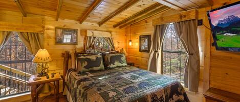 A cozy wooden cabin bedroom with a large bed, a small table with a lamp, and a TV on the wall.
