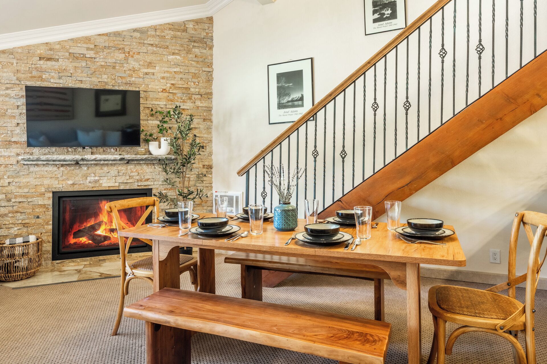 A dining area with a wooden table set for four, a fireplace, a television mounted on a brick wall, and a staircase with a wooden railing.