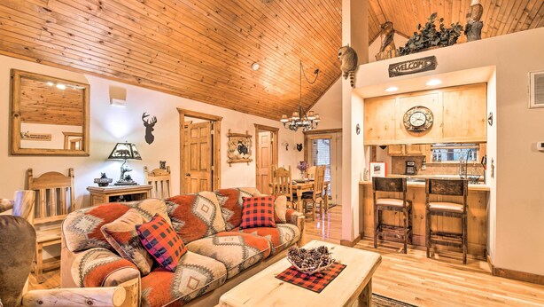 Een gezellige woonkamer met een houten plafond, een bank met opvallende kussens, een salontafel en een keukenhoek met houten kasten en een barstoel.
