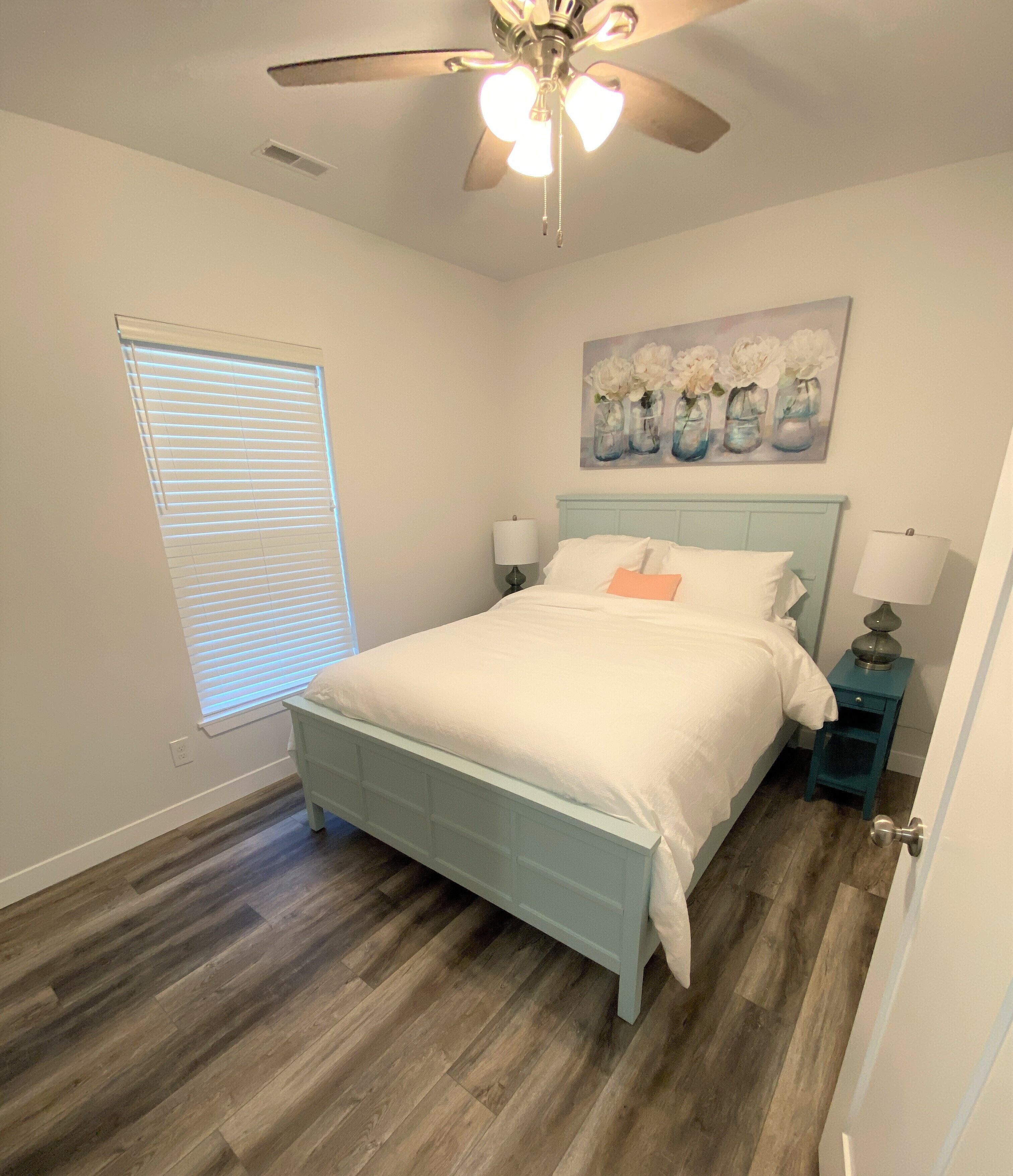 A bedroom with a bed, a ceiling fan, a window with blinds, a painting on the wall, and a nightstand with a lamp.