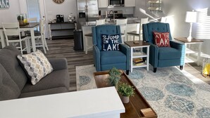 Un salon moderne avec un canapé gris, une table basse et deux fauteuils bleus ornés de coussins décoratifs. On aperçoit à l’arrière-plan un coin repas avec une table et des chaises blanches.