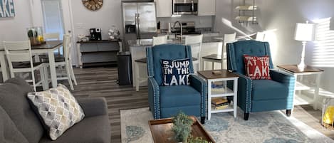 Una sala moderna con un sofá gris, una mesa de centro y dos sillones azules con cojines decorativos. Se ve una zona de comedor con una mesa y sillas blancas al fondo.