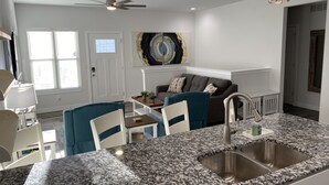Eine moderne Küche mit Granit Arbeitsplatten, einer Edelstahlspüle und einem Deckenventilator. Es gibt einen Essbereich mit Tisch und Stühlen, sowie ein Wohnzimmer mit einer Couch und einem Gemälde an der Wand.