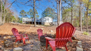 An outdoor seating area with red Adirondack chairs and a fire pit, surrounded by trees and a house in the background. - Waterfront Lake Hartwell Home w/ Dock & Fire Pit (Hartwell)