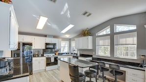 A modern kitchen with white cabinets, a large island with bar stools, and a view of the outdoors through large windows. - Waterfront Lake Hartwell Home w/ Dock & Fire Pit (Hartwell)